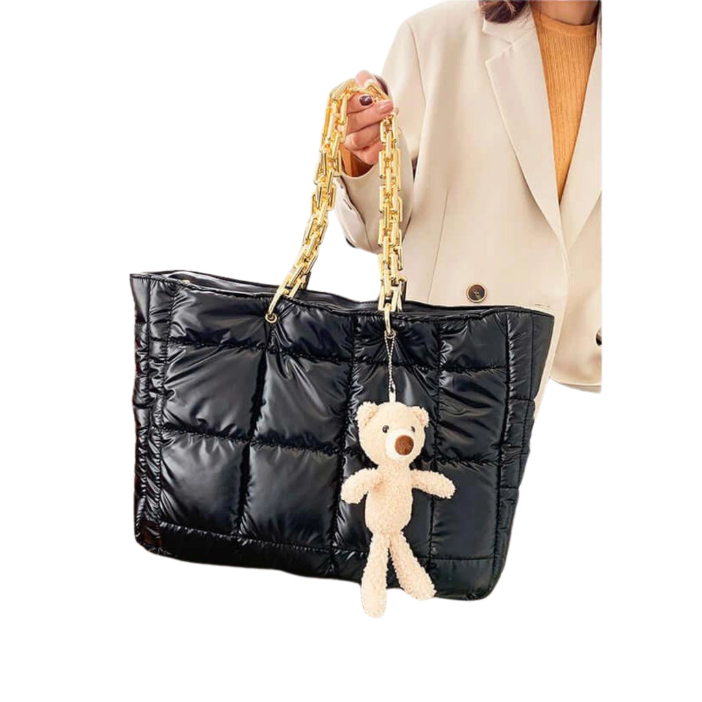 Schwarzer gesteppter Shopper mit Teddy-Anhänger und goldfarbener Kette vor heller Kleidung