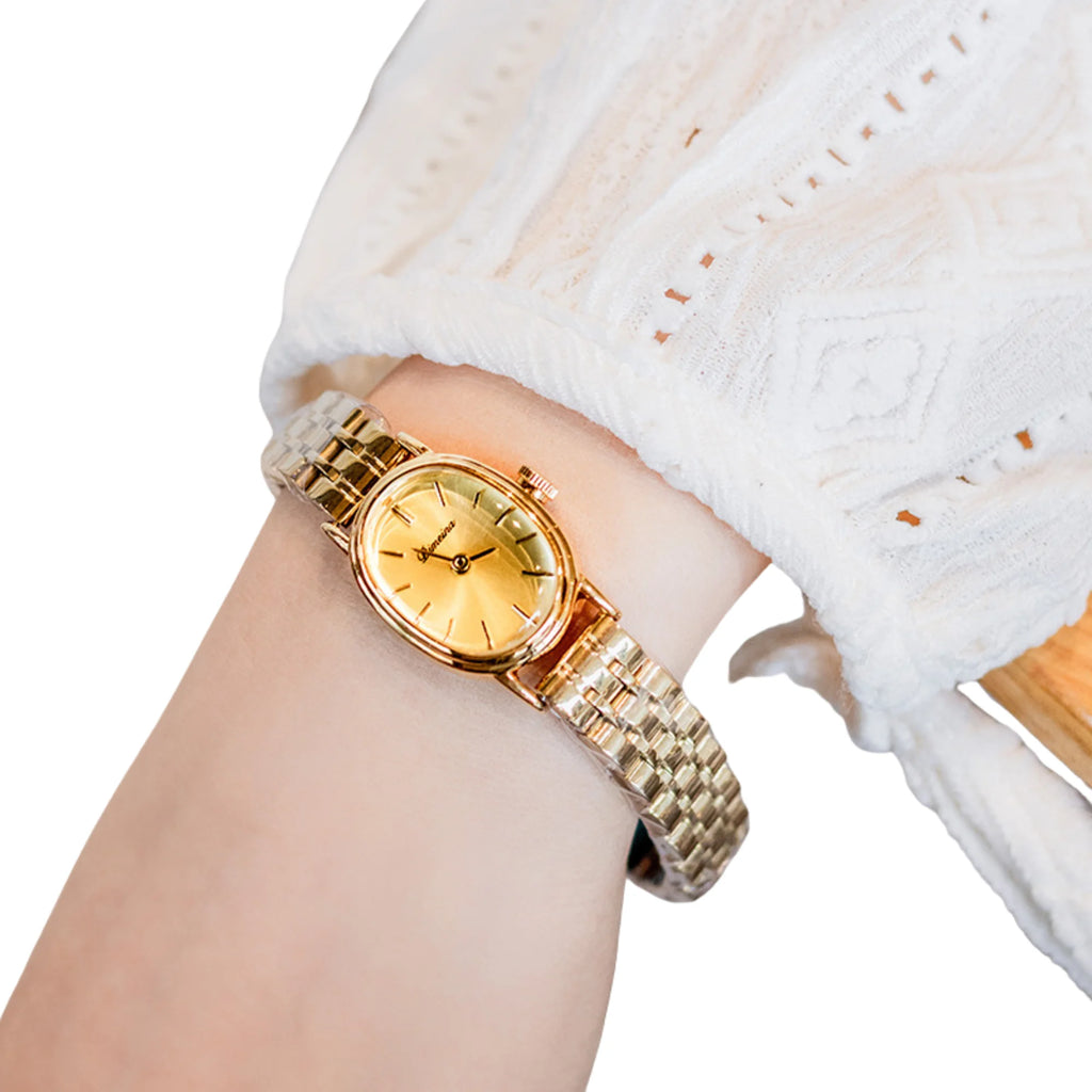 Goldene Uhr mit ovalem Gehäuse und elegantem Band, getragen mit weissem Spitzenärmel.