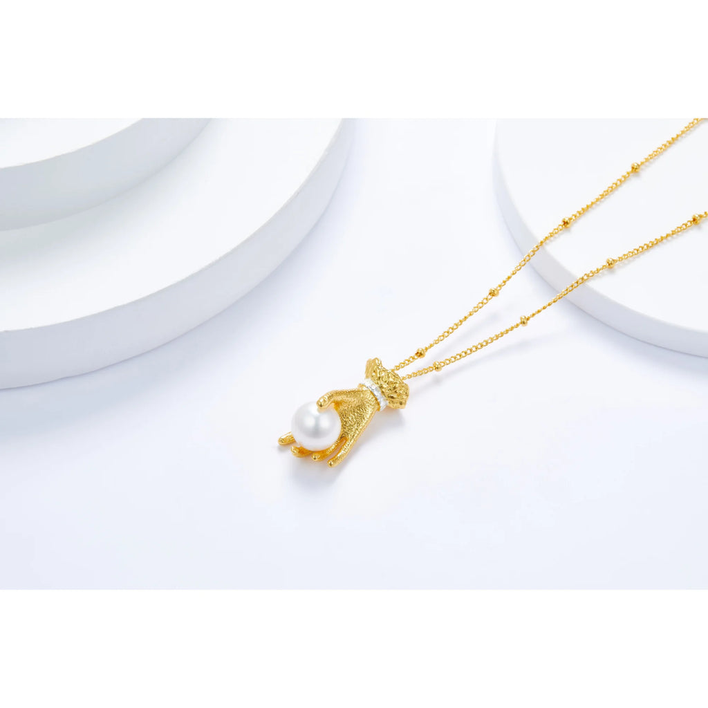 Skulpturale Kette mit vergoldetem Handanhänger, das eine Perle hält – abgelegt auf geometrischem weissem Objekt