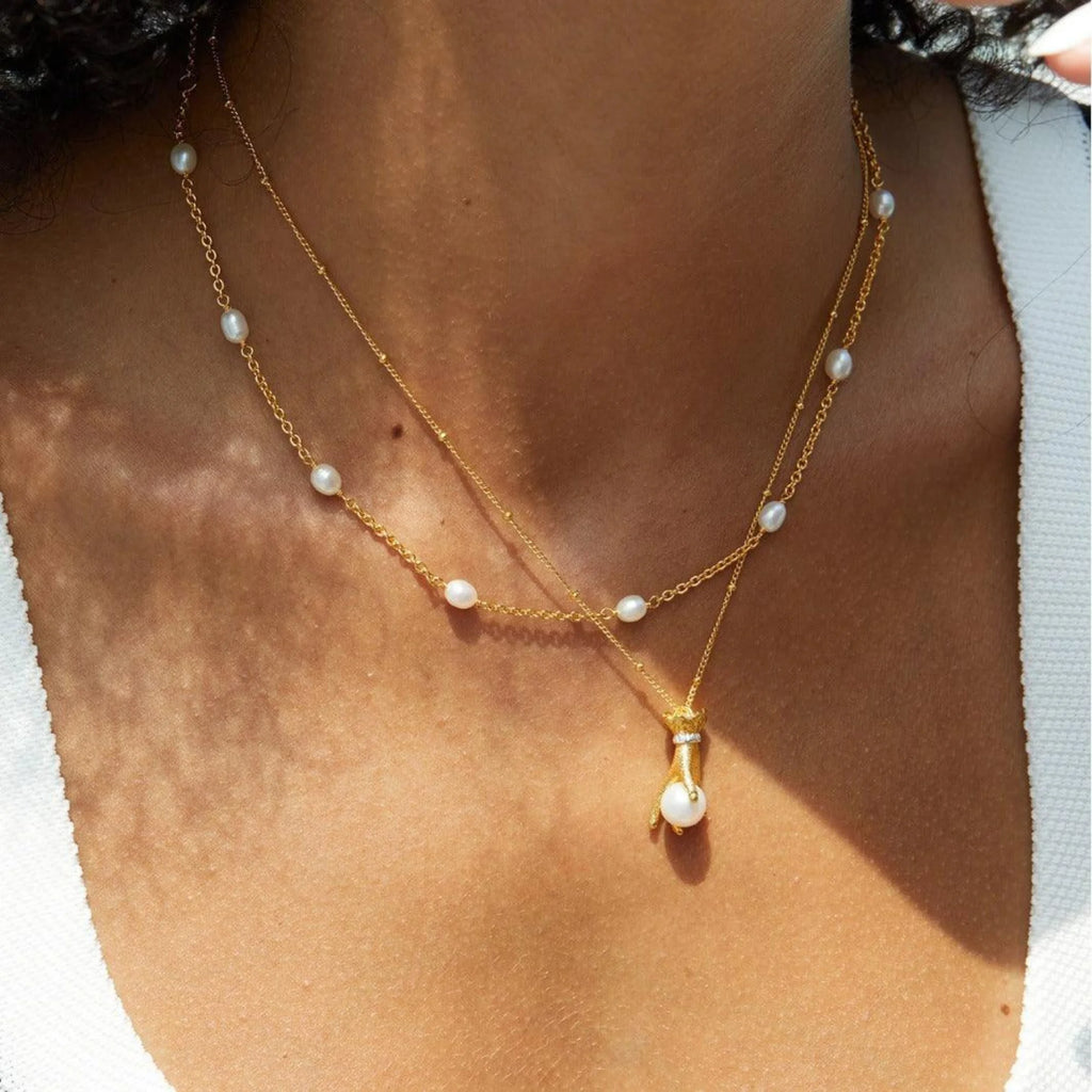 Zwei vergoldete Ketten im Layering-Look – obere Kette mit kleinen Perlen, untere mit einer goldenen Handform und Perlenanhänger