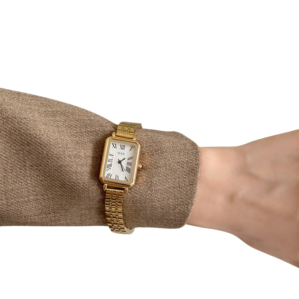  Goldene rechteckige Uhr mit römischen Ziffern am Handgelenk über braunem Langarmtop.