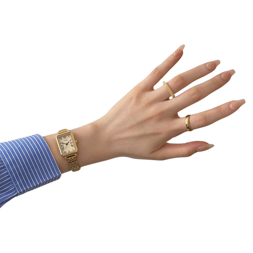 Goldene Uhr mit römischem Zifferblatt am Handgelenk mit schlichten Goldringen.