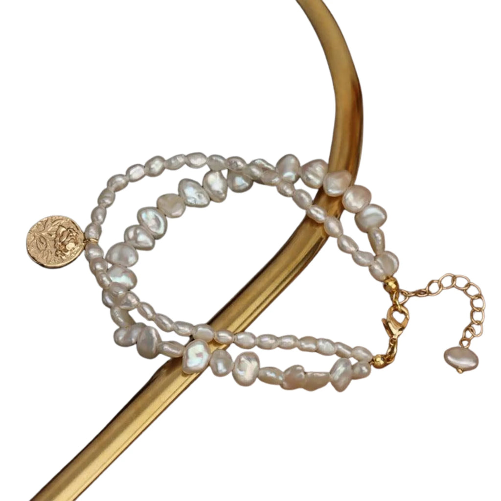 Zweireihiges Perlenarmband mit goldenem Coin-Anhänger – dekorativ über gebogenem Goldobjekt gelegt