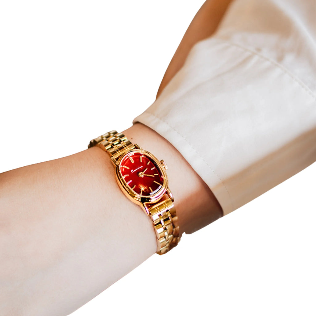 Goldene Uhr mit rotem Zifferblatt und Gliederarmband, getragen über einem beigen Ärmel.